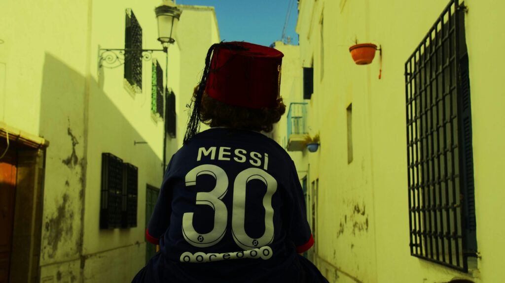 Back view of a person on a Tunis street wearing a fez and Messi jersey.