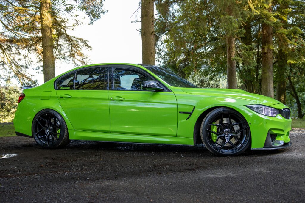 Green BMW M3 parked on a road amidst tall trees in England, showcasing luxury and performance.